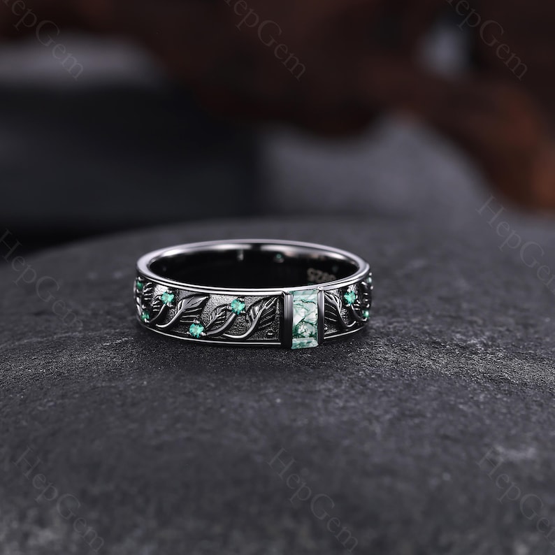 May include: A silver ring with a rectangular green stone at the center. The ring features a leaf and vine design with small green stones. The ring is set on a dark surface.