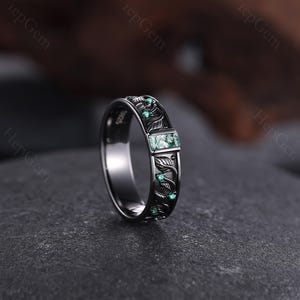 May include: A dark silver ring with an engraved leaf pattern and small green gemstones. The ring features a rectangular green stone at the center and is set on a dark gray surface. The ring is a piece of jewelry.