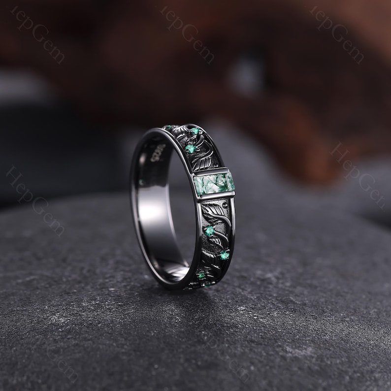 May include: A dark silver ring with an engraved leaf pattern and small green gemstones. The ring features a rectangular green stone at the center and is set on a dark gray surface. The ring is a piece of jewelry.