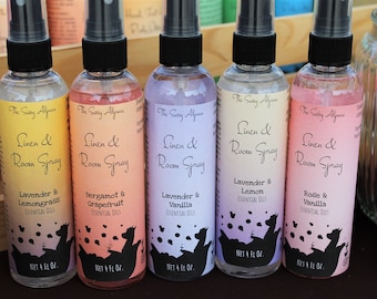 May include: Five clear glass spray bottles with black caps and labels. The labels are white with black text and feature a black silhouette of a dog. The text on the labels reads "The Sally Alyce Linen & Room Spray" and lists different essential oil scents: Lavender & Lemongrass, Bergamot & Grapefruit, Lavender & Vanilla, Lavender & Lemon, and Rose & Vanilla. Each bottle contains 4 fluid ounces.