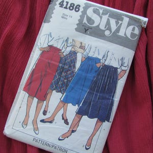 Puede incluir: Patrón de costura vintage titulado "Style" con ilustraciones de faldas. El patrón es talla 14 Miss, con un diseño blanco y azul. El fondo es una tela roja oscura texturizada.