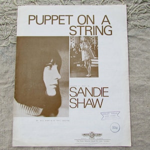 May include: A vintage sheet music cover for the song "Puppet on a String" by Sandie Shaw. The cover features a black and white photo of Sandie Shaw, a woman with long hair, looking to the side. The cover is printed on a light brown background with the title in bold, black letters.
