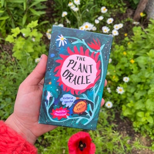 May include: A hand holds a deck of cards called "The Plant Oracle" with a colourful illustration of a flower and the text "Victoria Dorche, Mathilde Morisset, Diane Arriché".