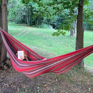 Può includere: Un'amaca a righe rosse e bianche è appesa tra due alberi in un'area erbosa. Una persona si rilassa nell'amaca, leggendo un libro. L'amaca è fatta di tessuto intrecciato.