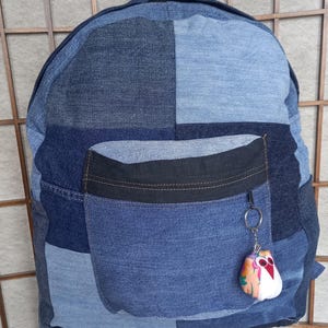 Può includere: Uno zaino in denim blu con una grande tasca frontale e un piccolo portachiavi a forma di gufo. Lo zaino è realizzato in denim blu di diverse tonalità e ha una cerniera nera sulla tasca frontale.