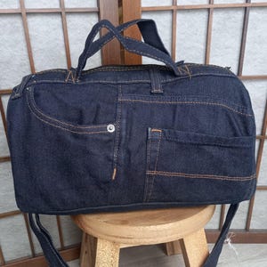 Op de afbeelding: Een donkerblauwe denim duffeltas met oranje stiksels. De tas heeft een ritssluiting, twee handvatten en een schouderriem. Het ontwerp bevat jeans-elementen, waaronder zakken en klinknagels.