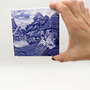 May include: A square, blue and white ceramic tile with a detailed village scene illustration. The tile shows buildings, trees, and figures in a traditional style. The word "Madeira" is visible on the tile.