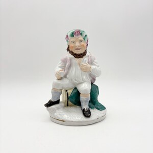 Peut inclure: Figurine en porcelaine vintage d'un homme assis, vêtu d'une chemise blanche, d'un pantalon et de chaussures noires. La figurine a une barbe brune et un couvre-chef floral. L'homme est assis sur un tissu vert, tenant une petite tasse.