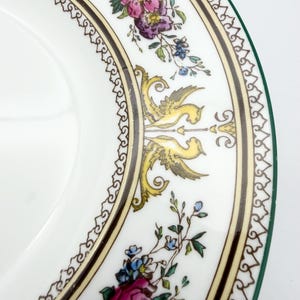 May include: White ceramic plate with a decorative floral and bird design. The plate features a border with a repeating pattern, and a green edge. The floral design includes pink, purple, and blue flowers, with yellow and gray birds.
