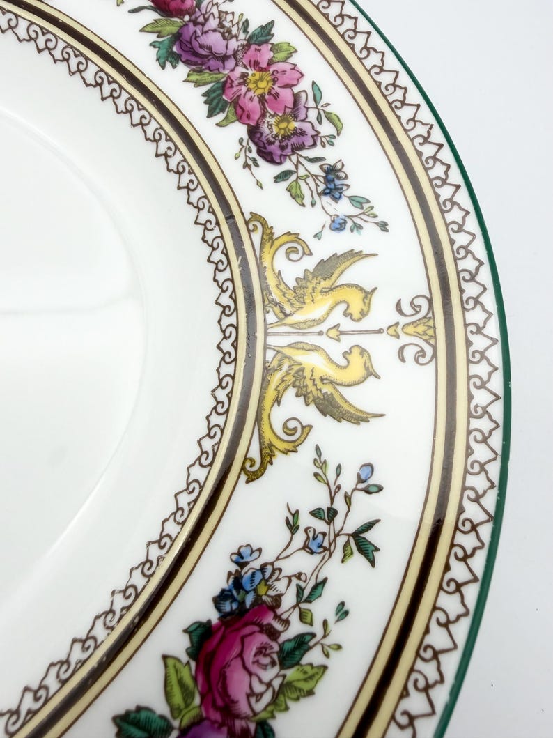 May include: White ceramic plate with a decorative floral and bird design. The plate features a border with a repeating pattern, and a green edge. The floral design includes pink, purple, and blue flowers, with yellow and gray birds.