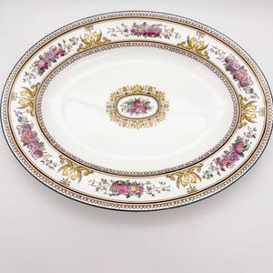 May include: Oval serving platter with a white center and a decorative border. The border features pink and purple floral arrangements, gold accents, and a brown trim. The platter has a green edge and a central floral design.