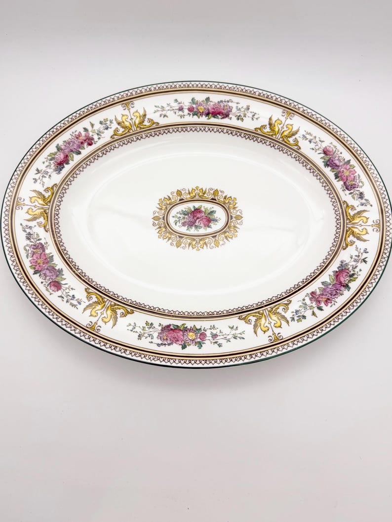 May include: Oval serving platter with a white center and a decorative border. The border features pink and purple floral arrangements, gold accents, and a brown trim. The platter has a green edge and a central floral design.