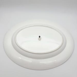 May include: White oval serving platter with a raised rim. The underside of the platter is visible, featuring a small emblem and text. The platter is likely made of ceramic or porcelain, suitable for serving food.