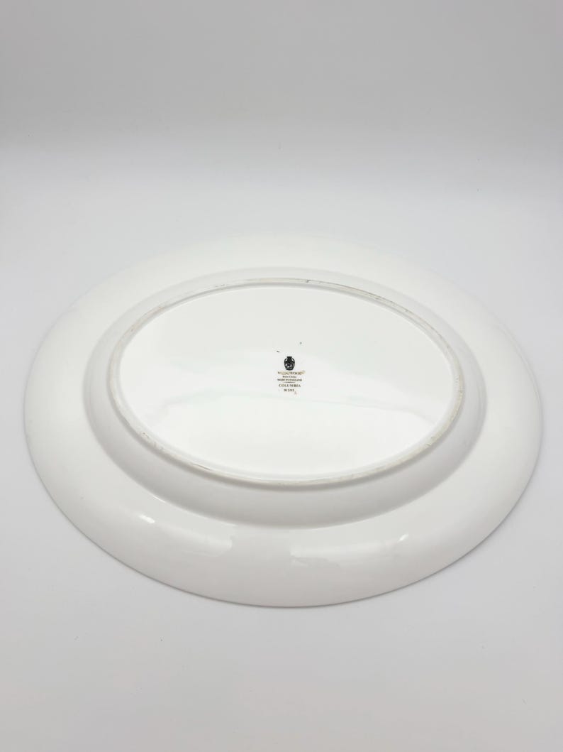 May include: White oval serving platter with a raised rim. The underside of the platter is visible, featuring a small emblem and text. The platter is likely made of ceramic or porcelain, suitable for serving food.
