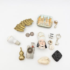 May include: A selection of antique objects: a gold-toned leaf brooch, a small tin with a building illustration, a miniature camera, dice, and a key charm. Other items include a chef figurine, a gourd-shaped object, and a white pipe.