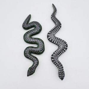 Puede incluir: Dos esculturas decorativas de serpientes. Una es negra con patrones de puntos blancos y verdes, la otra es negra con patrones de puntos blancos. Ambas tienen una forma curva y serpentina, sobre un fondo blanco.