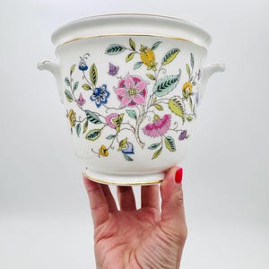 May include: A white ceramic planter with a floral design. The planter features a colourful pattern of pink, blue, yellow, and green flowers and leaves. It has a gold trim and two small handles. The planter is held up by a hand.