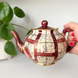 May include: A ceramic teapot featuring a cream-coloured base with a dark red and gold plaid pattern, overlaid with gold floral line art. The teapot has a matching lid with a gold rim, a curved spout, and a handle, with the text Royal Bradwell.