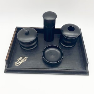May include: A black wooden tray with a silver letter "P" on the front. The tray contains four black wooden containers, including a round dish, a cylindrical jar, a small round jar with a lid, and a taller cylindrical jar with a lid.