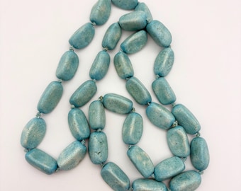 Op de afbeelding: Een ketting met ovale, blauwgroene kralen. De kralen zijn geregen met een bijpassend blauwgroen koord, wat een eenvoudig ontwerp creëert. De ketting wordt gepresenteerd tegen een witte achtergrond. De kralen zijn ongeveer 1,5 cm lang.