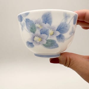 K&ouml;nnte beinhalten: Eine wei&szlig;e Keramik-Teetasse mit floralem Design. Die Tasse hat blaue und gr&uuml;ne Blumen und Bl&auml;tter. Der Boden der Tasse hat blaue Streifen. Die Tasse wird von einer Person gehalten.