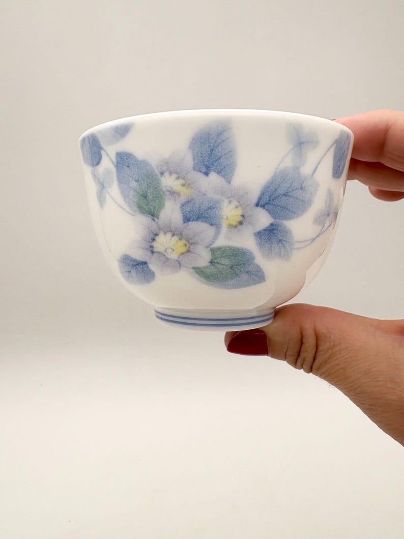 K&ouml;nnte beinhalten: Eine wei&szlig;e Keramik-Teetasse mit floralem Design. Die Tasse hat blaue und gr&uuml;ne Blumen und Bl&auml;tter. Der Boden der Tasse hat blaue Streifen. Die Tasse wird von einer Person gehalten.