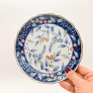Puede incluir: Un plato redondo de cerámica con borde festoneado. El plato presenta un borde azul con un diseño floral y un centro blanco con hojas azules y bayas naranjas. El plato está sostenido por una mano.