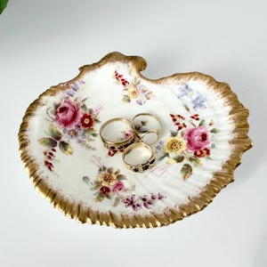 May include: A white porcelain shell-shaped trinket dish with a gold-painted scalloped edge, decorated with floral patterns of pink roses and yellow flowers, containing three gold rings resting in the center of the dish.