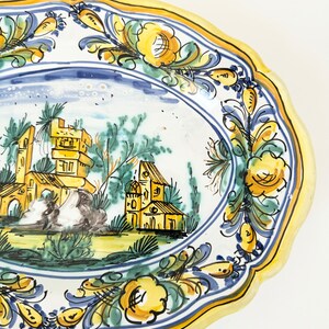 Pu&ograve; includere: Un piatto ovale in ceramica giallo e blu con un disegno dipinto a mano di una scena di paese con edifici e alberi. Il piatto ha un bordo ondulato e un bordo floreale.