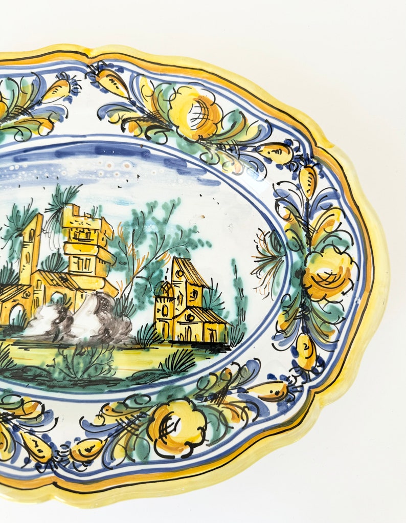 Pu&ograve; includere: Un piatto ovale in ceramica giallo e blu con un disegno dipinto a mano di una scena di paese con edifici e alberi. Il piatto ha un bordo ondulato e un bordo floreale.