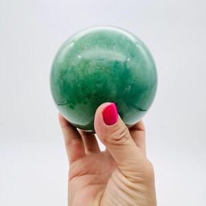 May include: A green sphere with a mottled texture, held in a hand with bright pink nail polish. The sphere appears to be made of stone or a similar material. The background is plain white. The sphere's diameter is approximately 7 cm.
