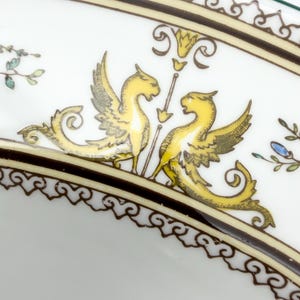 May include: White ceramic plate with a decorative border and a central design featuring two yellow winged creatures facing each other. The plate has a brown and gold border with ornate scrollwork and floral accents.