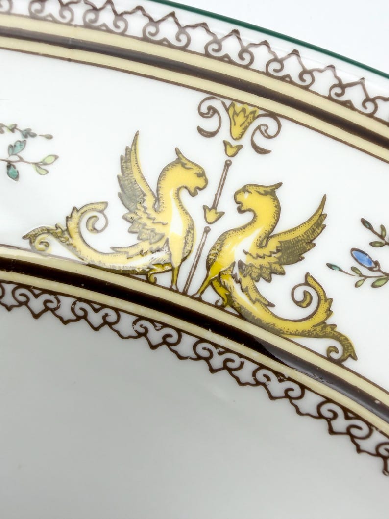 May include: White ceramic plate with a decorative border and a central design featuring two yellow winged creatures facing each other. The plate has a brown and gold border with ornate scrollwork and floral accents.