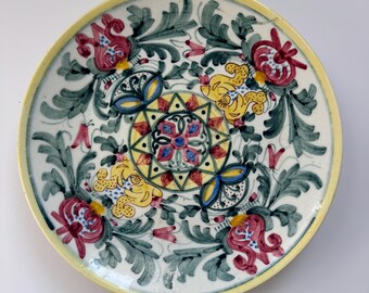 May include: A hand-painted ceramic plate with a yellow rim and intricate floral designs. The plate features red, yellow, and blue flowers with green leaves, set against a white background. The center has a geometric pattern.
