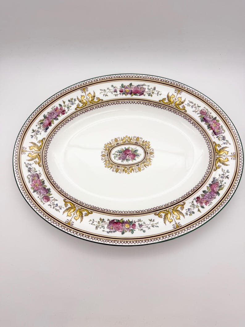 May include: Oval serving platter with a white background. The platter features a floral design with pink and purple flowers, gold accents, and a brown border. The rim is green.
