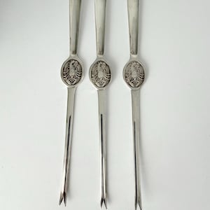 May include: Three silver-toned metal seafood forks arranged vertically. Each fork features a smooth, elongated handle and a two-pronged tip. An oval medallion is centered on each handle, displaying a detailed embossed lobster design in relief.