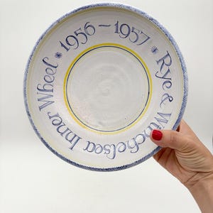 Könnte beinhalten: Ein weißer Keramikteller mit blauem und gelbem Rand, mit blauem Text, der "Wheel Inner Chelsea 1956-1957 Rye & Winch" lautet. Der Teller hat ein handgemaltes Aussehen.