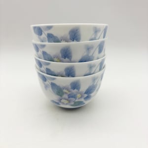 K&ouml;nnte beinhalten: Ein Stapel von vier wei&szlig;en Keramikschalen. Jede Schale ist mit einem zarten Blumendesign mit blauen Bl&auml;ttern und hellvioletten Bl&uuml;ten verziert. Die Schalen sind sauber gestapelt und zeigen das filigrane Muster.