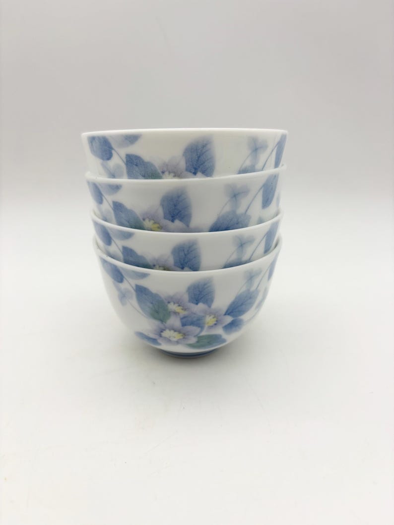K&ouml;nnte beinhalten: Ein Stapel von vier wei&szlig;en Keramikschalen. Jede Schale ist mit einem zarten Blumendesign mit blauen Bl&auml;ttern und hellvioletten Bl&uuml;ten verziert. Die Schalen sind sauber gestapelt und zeigen das filigrane Muster.