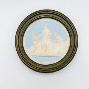 Puede incluir: Una placa de pared circular con fondo azul claro y un relieve blanco de una figura central sentada entre dos figuras más pequeñas, una de las cuales sostiene una corona. La pieza está encerrada en un marco metálico circular oscuro y desgastado.