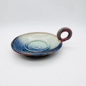 May include: Handmade ceramic dish with a spiral design in the center. The dish features a blue, white, and red color scheme, with a circular handle. The dish is approximately 6 inches in diameter.