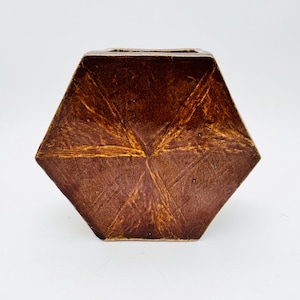 May include: A brown, hexagonal vase with a textured surface. The vase has a wide, open top and a geometric design. The color is a rich, earthy brown with lighter, golden-brown accents.