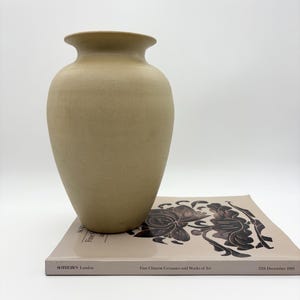 May include: A beige ceramic vase with a wide base and a flared rim, placed on a book. The book features a floral design and the text "SOTHEBY'S London" and "Fine Chinese Ceramics and Works of Art", dated "12th December 1989".
