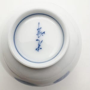 K&ouml;nnte beinhalten: Eine wei&szlig;e Keramikschale mit blauem Rand und blauem Anstrich. Der Boden der Schale zeigt blaue japanische Kalligraphie. Die Schale ist rund und hat eine glatte, gl&auml;nzende Oberfl&auml;che.