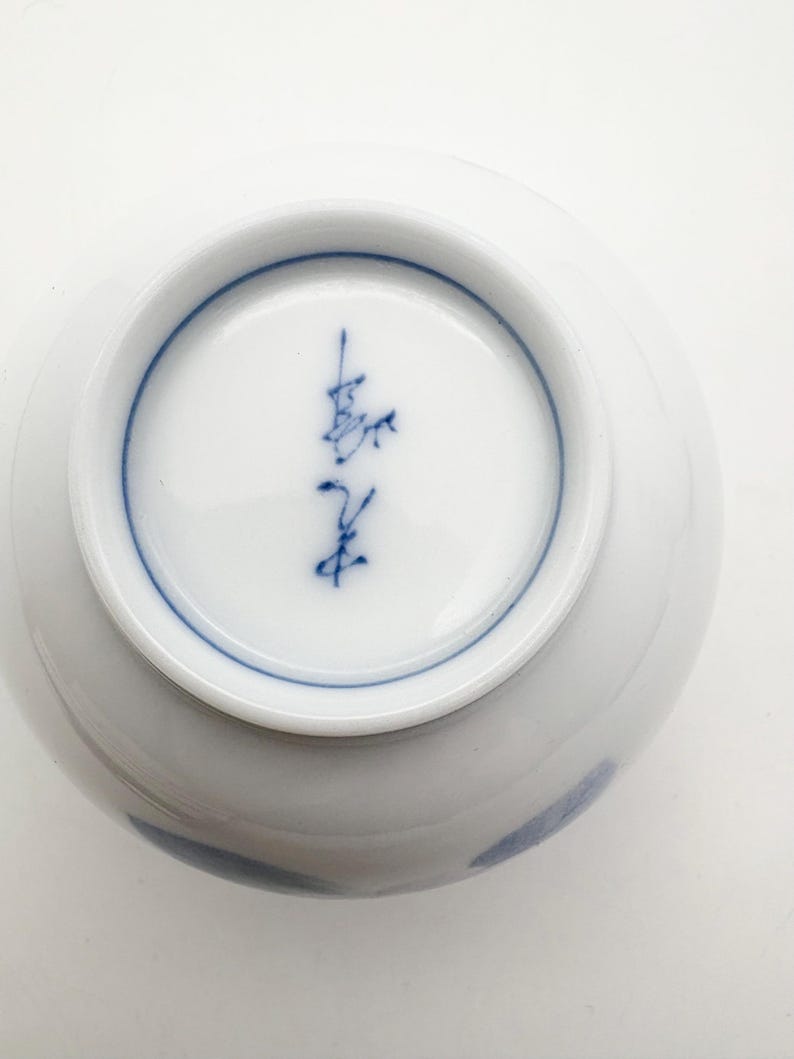 K&ouml;nnte beinhalten: Eine wei&szlig;e Keramikschale mit blauem Rand und blauem Anstrich. Der Boden der Schale zeigt blaue japanische Kalligraphie. Die Schale ist rund und hat eine glatte, gl&auml;nzende Oberfl&auml;che.