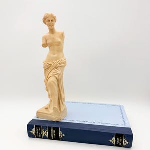 Puede incluir: Una estatua beige de Venus de Milo se encuentra sobre un libro azul marino. La estatua es una representación clásica de la diosa, con tela drapeada y brazos faltantes. El libro tiene letras doradas y una cubierta azul claro.