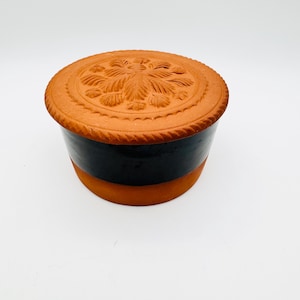 May include: A terracotta pottery butter dish with a black glazed band around the middle. The lid has a carved floral design.