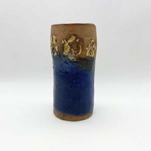 Puede incluir: Un jarrón de cerámica hecho a mano con un esmalte azul y detalles marrones. El jarrón tiene una forma cilíndrica simple y está decorado con tres diseños de flores blancas.