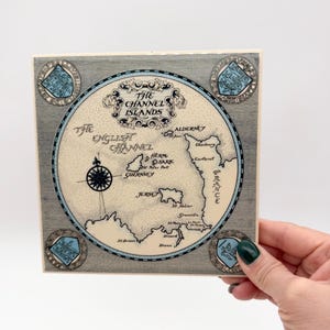 May include: Square ceramic tile featuring a map of the Channel Islands, with the text "The Channel Islands" and "The English Channel". The map is in shades of blue, beige, and gray, with decorative elements and a compass rose. Four crests are in each corner.