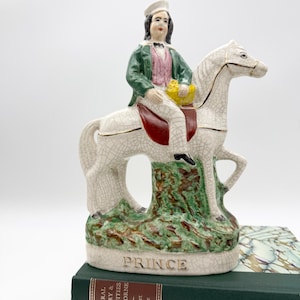 Könnte beinhalten: Vintage Keramikfigur einer Person, die ein weißes Pferd reitet. Der Reiter trägt eine grüne Jacke, eine rosa Weste und einen weißen Hut. Der Sockel ist grün und braun mit dem Wort "PRINCE" in Weiß. Die Figur steht auf einem grünen Buch.